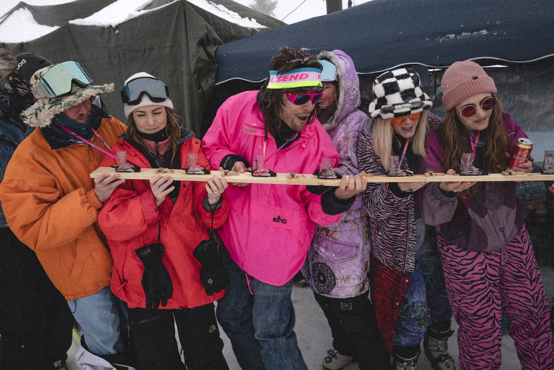 Gruppe in bunten Retro-Skianzügen stößt gemeinsam am langen Holz-Shotski an