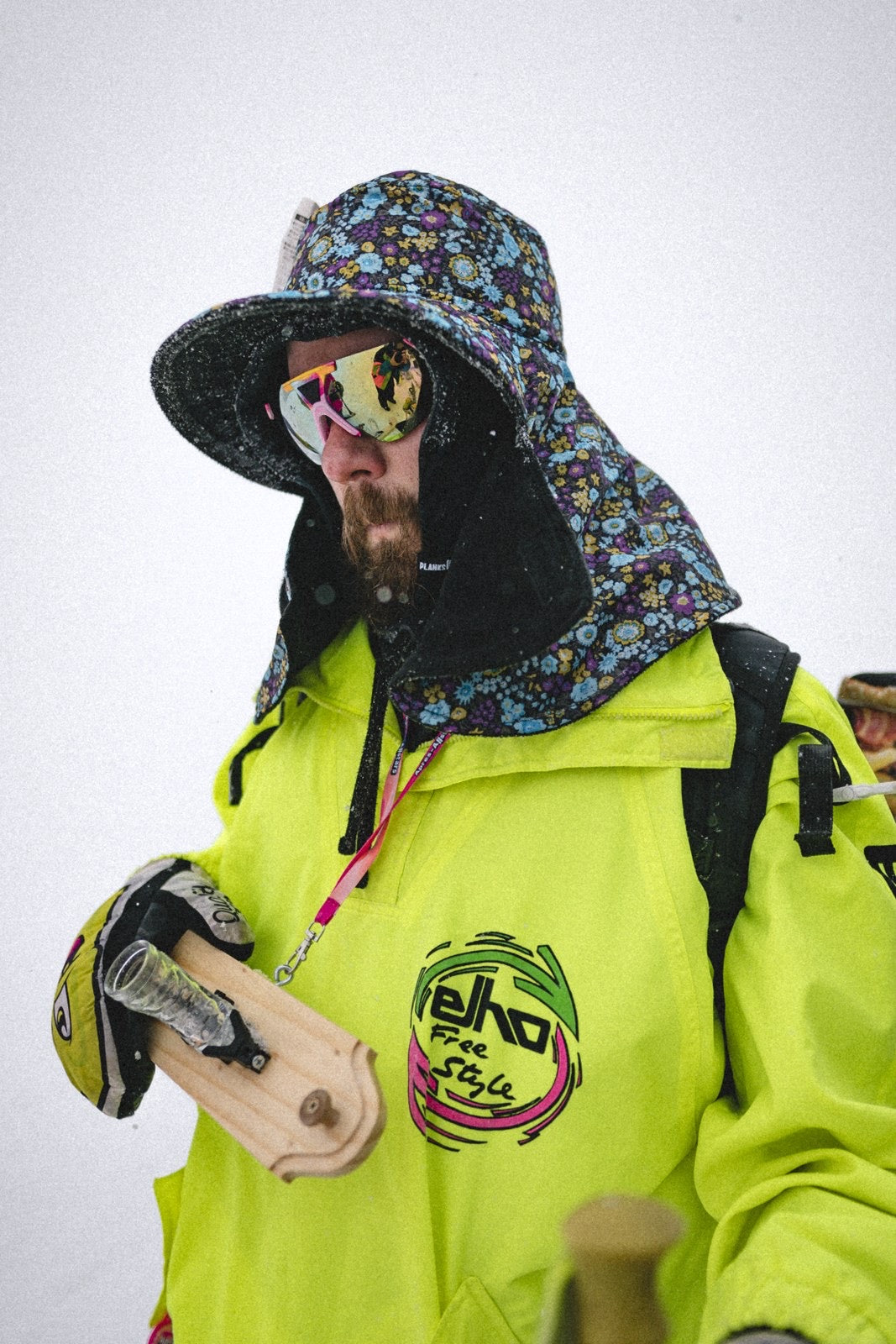 Freerider in Neonjacke und Bucket Hat hält einen Holz-Shotski mit Skischuh-Shotglas im Schnee
