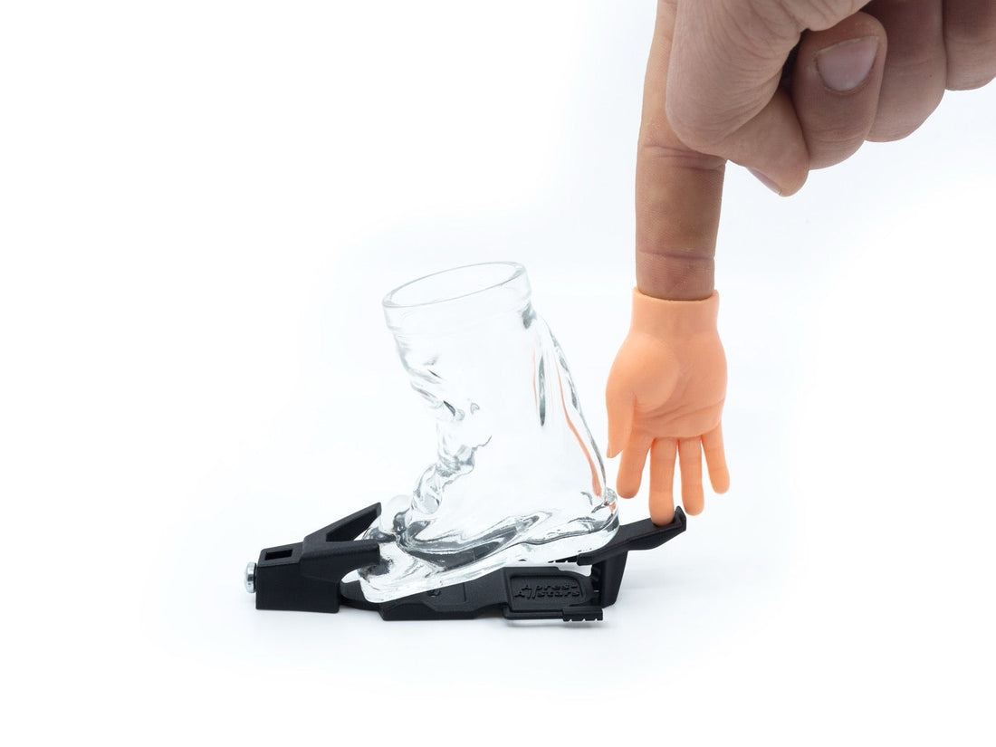 Minihand auf einer Fingerkuppe neben einem Skischuh-Shotglas mit Bindung